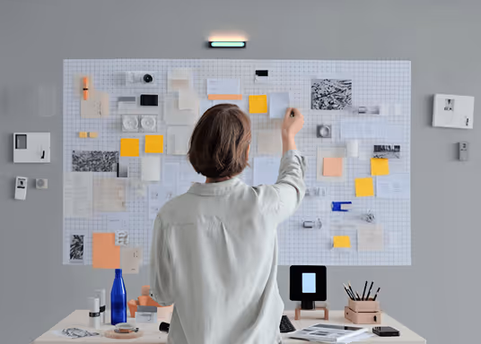 Person with short brown hair facing a wall covered with a large grid paper and various sticky notes and papers, standing next to a desk with office supplies.