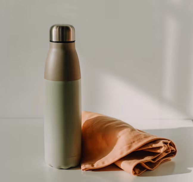 Matte metallic water bottle next to a folded light brown cloth on a white surface with soft natural light.